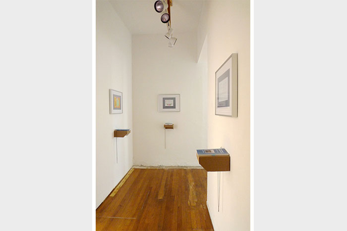 documentation photo of a narrow hallway containing three framed photographs and three books atop cardboard shelves underneath each framed photo