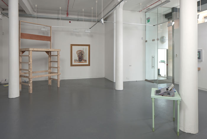 documentation photo of a room with three pillars containing a large wooden platform, a large framed photograph, and a green metal desk with stacks of booklets and a clear plastic typewriter on top of it